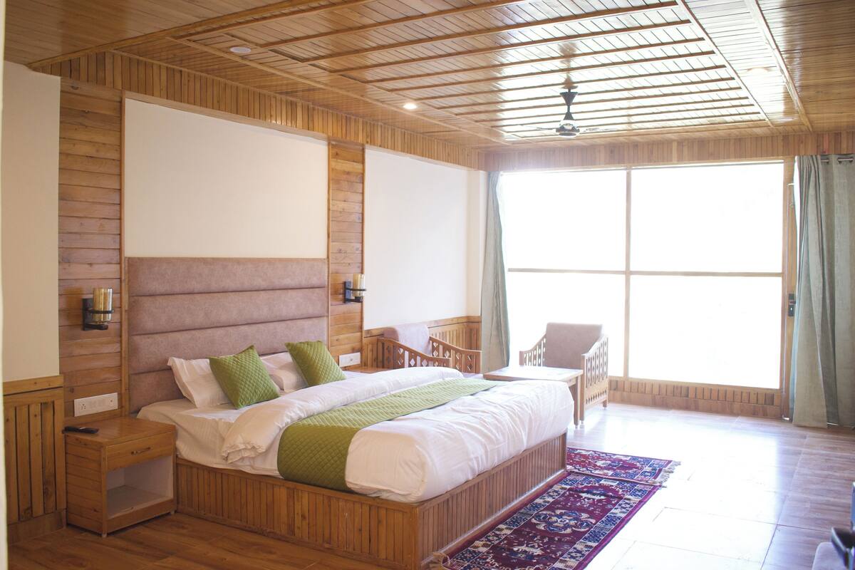 A spacious bedroom with a large bed, wooden headboard, bedside tables, and a view of the outdoors through a large window.