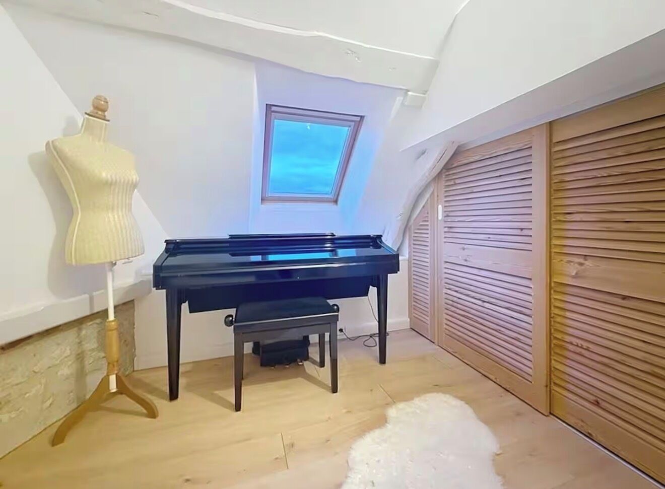 Un piano dans une pièce dont le plafond est en pente, un mannequin, une lampe et une vue à travers une lucarne.
