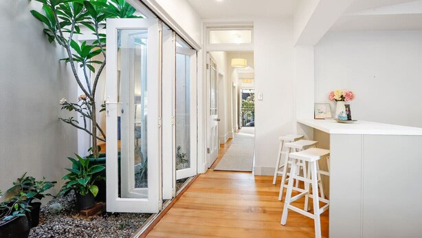 Un pasillo moderno con suelo de madera, paredes blancas y un vidrio que da a un balcón. Hay plantas en macetas y una pequeña zona de bar con taburetes.