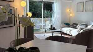 Un salon moderne avec un canapé blanc, une table à manger agrémentée de vases et une vue sur l’extérieur grâce à de grandes fenêtres.