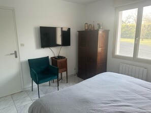 A bedroom with a bed, a television mounted on the wall, a wooden cabinet, and a window with a view of greenery. - Le Petit Chatelier - Detached Country House With Large Garden in the Loire Valley (Mauges-sur-Loire)