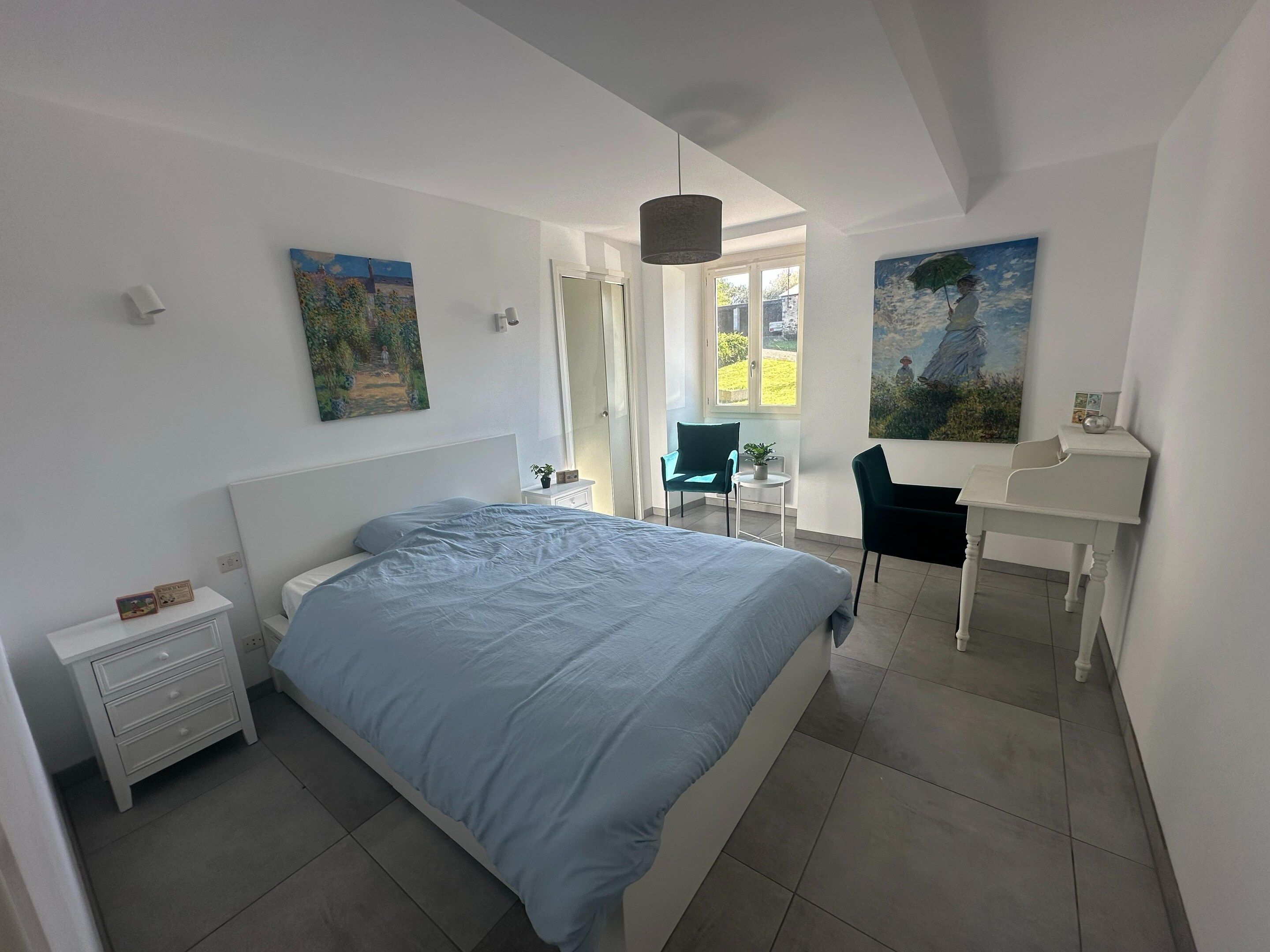 A bedroom with a bed, a desk, and two chairs. There are paintings on the walls and a window providing natural light.