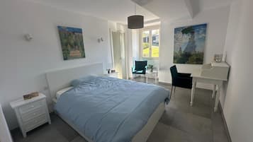 A bedroom with a bed, a desk, and two chairs. There are paintings on the walls and a window providing natural light.