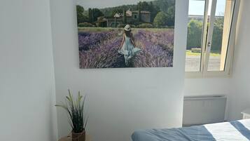 A bedroom with a bed, a window, a wall-mounted painting of a woman in a field, and a small round table with a plant.