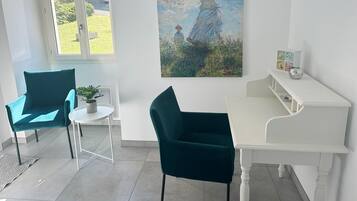 A room with a white desk, two teal chairs, a small table with a plant, and a painting of a woman with an umbrella.