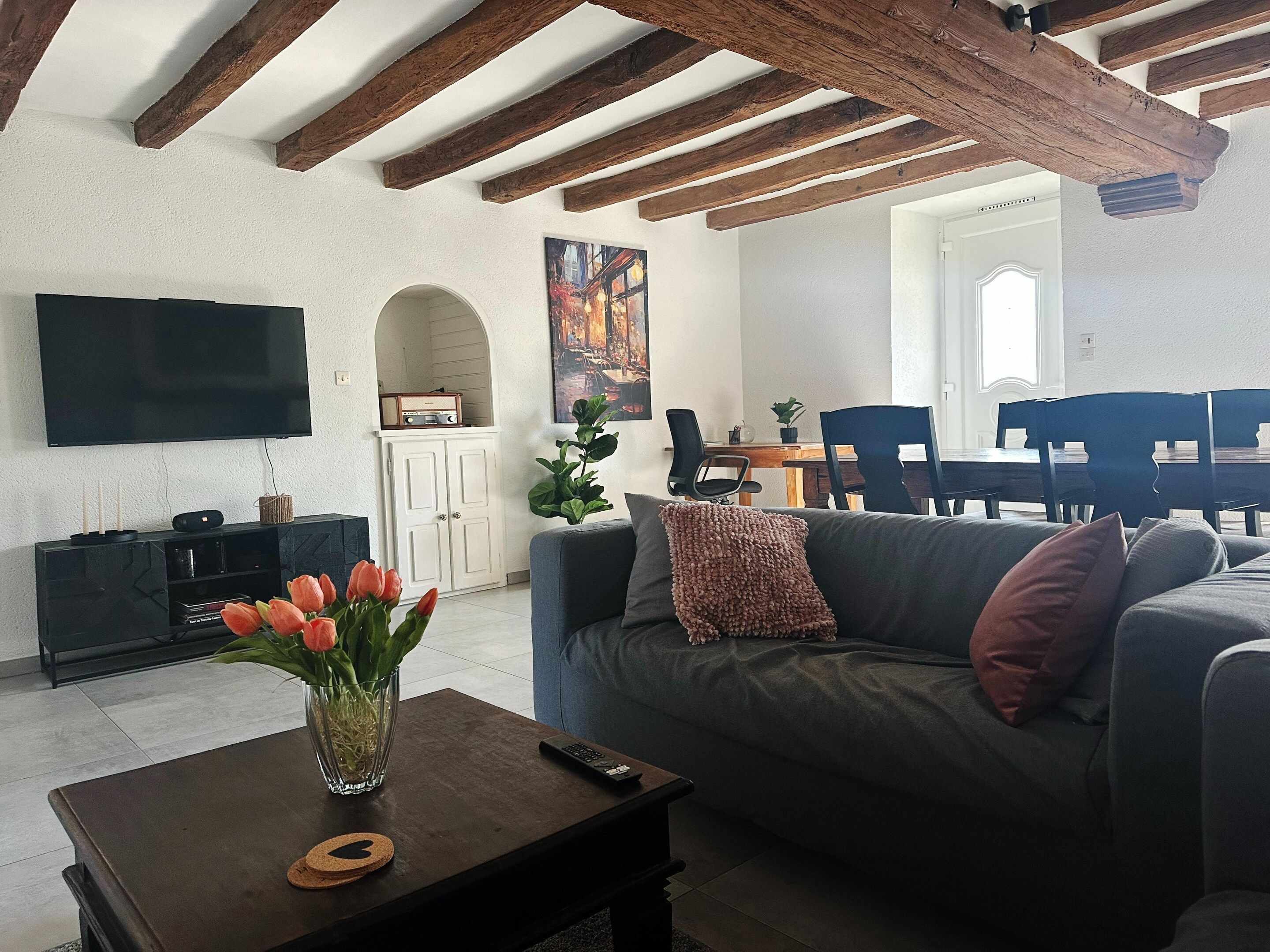A living room with a sofa, a coffee table with a vase of flowers, a flat-screen TV, and a dining area with chairs and a table.