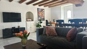 A living room with a sofa, a coffee table with a vase of flowers, a flat-screen TV, and a dining area with chairs and a table.