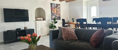 A living room with a sofa, a coffee table with a vase of flowers, a flat-screen TV, and a dining area with chairs and a table.