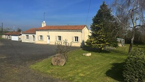 Un bâtiment avec un toit recouvert de tuiles, une cheminée et un petit jardin agrémenté d’une pierre, d’un arbre et d’un arbuste.