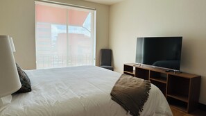 A hotel room with a large bed, a television on a wooden stand, a chair, and a balcony with a view of buildings. - Lux 2-BR 1 story Condo in the heart of Everything!!! (San Rafael de Escazú)