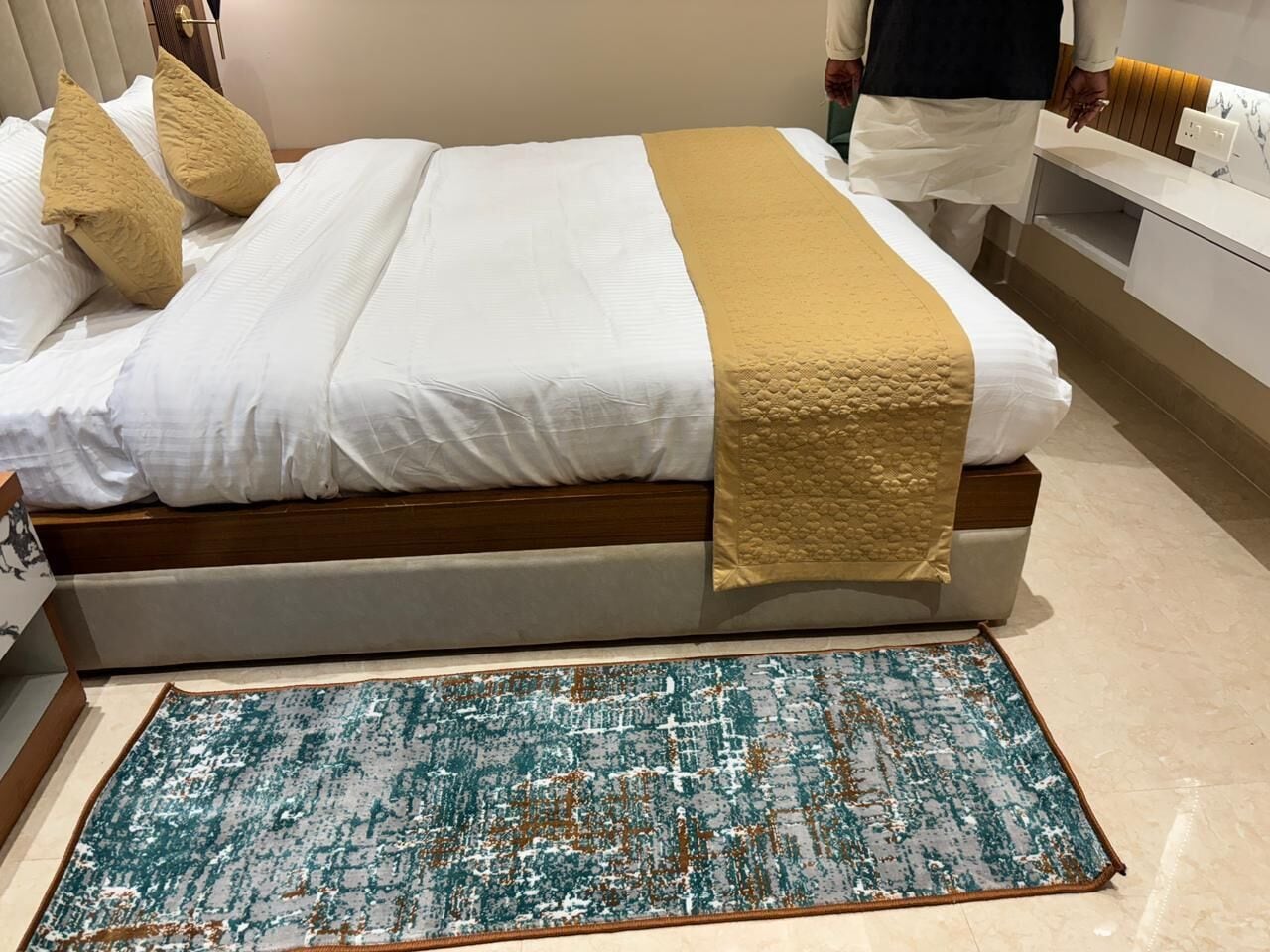 Una cama con cubrecama blanco y beige, una manta dorada y dos almohadas beige. Un tapete azul y blanco con aspecto desgastado se encuentra frente a la cama.