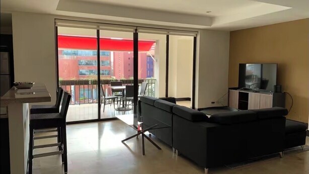 A modern living room with a sofa, a TV, and a dining area with a table and chairs. - Gorgeous 3-BR Condo in the heart of everything! (San Rafael de Escazú)