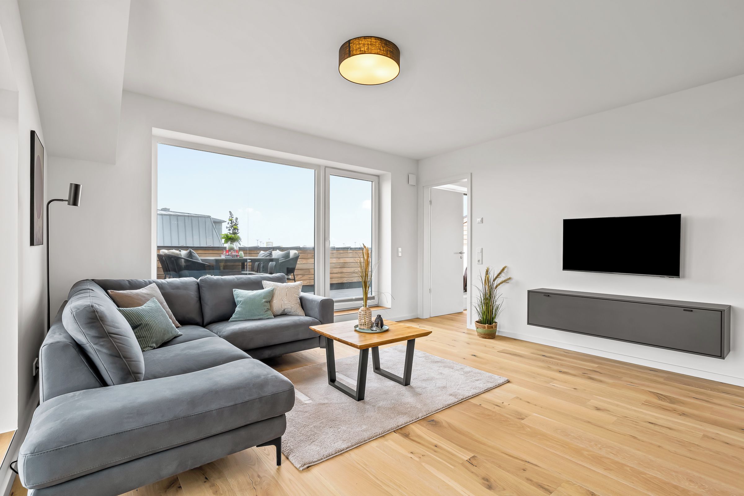 A modern living room with a grey sectional sofa, a wooden coffee table, and a flat-screen TV mounted on the wall.