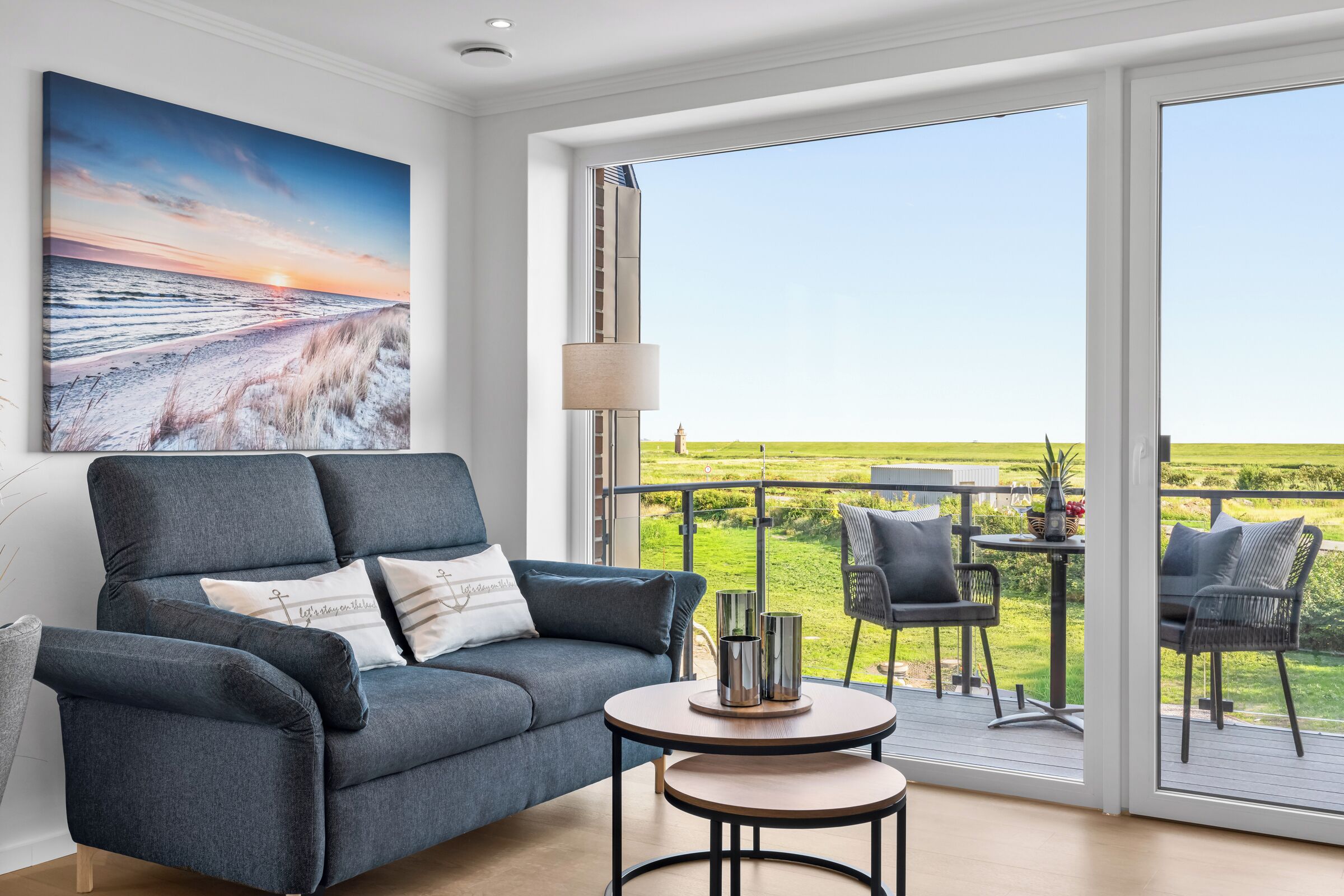 Ein modernes Wohnzimmer mit einem grauen Sofa, einem kleinen runden Tisch und Blick auf ein grünes Feld durch große Schiebetüren.