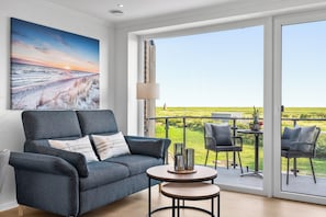 A modern living room with a grey sofa, a small round table, and a view of a green field through large sliding doors. - The View G02 Perle - Sea view, balcony, garden furniture,1st floor,parking space (Dagebüll)