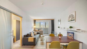 A modern living room with a sofa, coffee table, and dining table with chairs. There is a window with curtains, a radiator, and a door leading to another room.