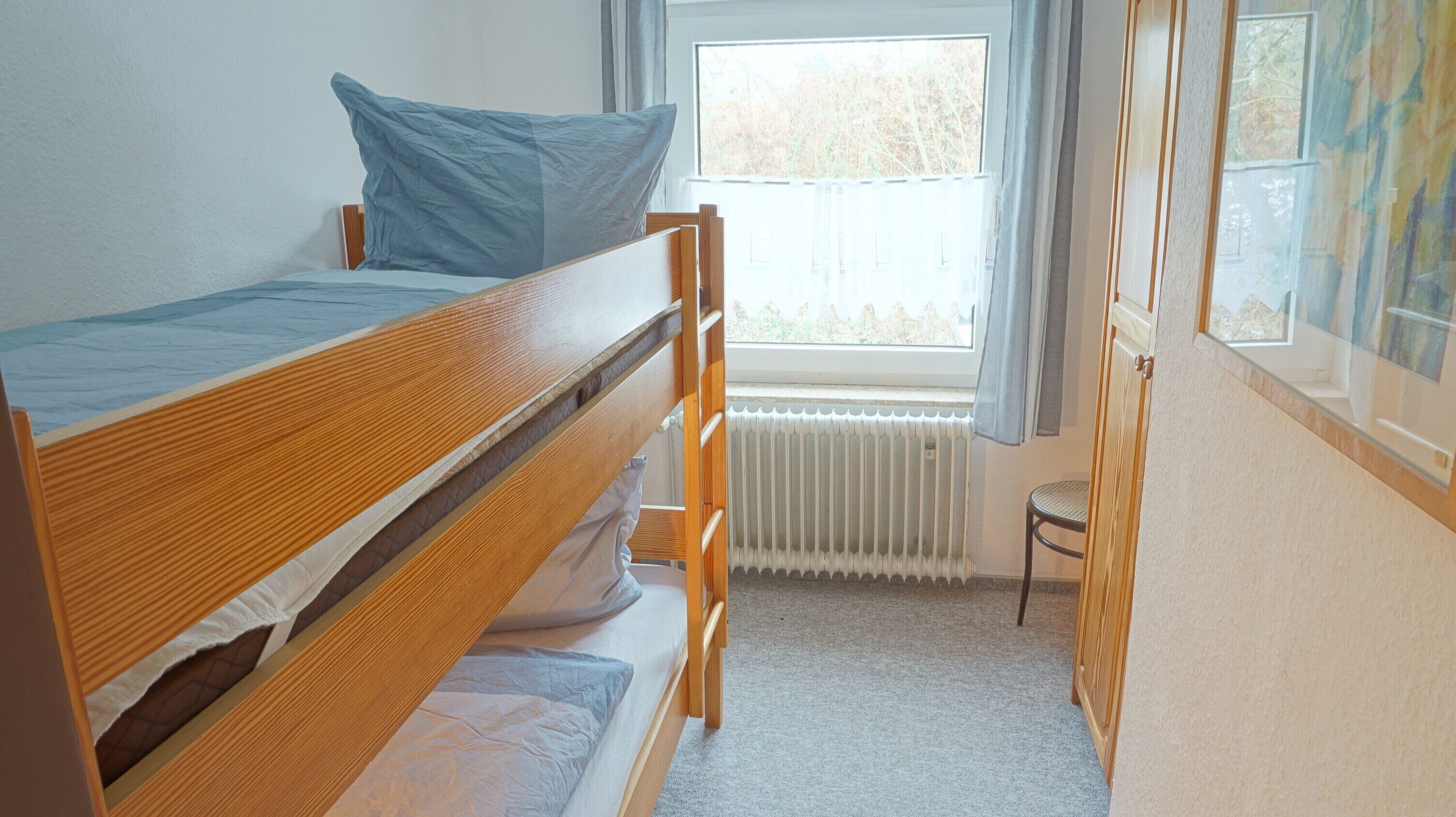 A small, single-bed room with a wooden bunk bed, a radiator, a window with curtains, and a chair.
