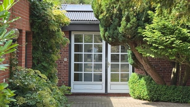 A brick building with a white door and windows, surrounded by lush greenery and trees. - Comfortable vacation apartment with terrace and family facilities in Garding (Garding)