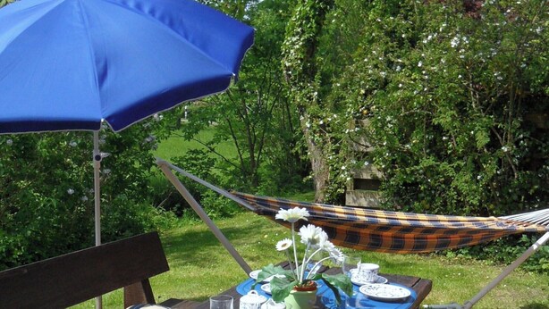 A picnic setup with a table, chairs, a hammock, and a blue umbrella. - Künstlergarten Apartment 2: Comfortable vacation accommodation with terrace and garden i (Garding)