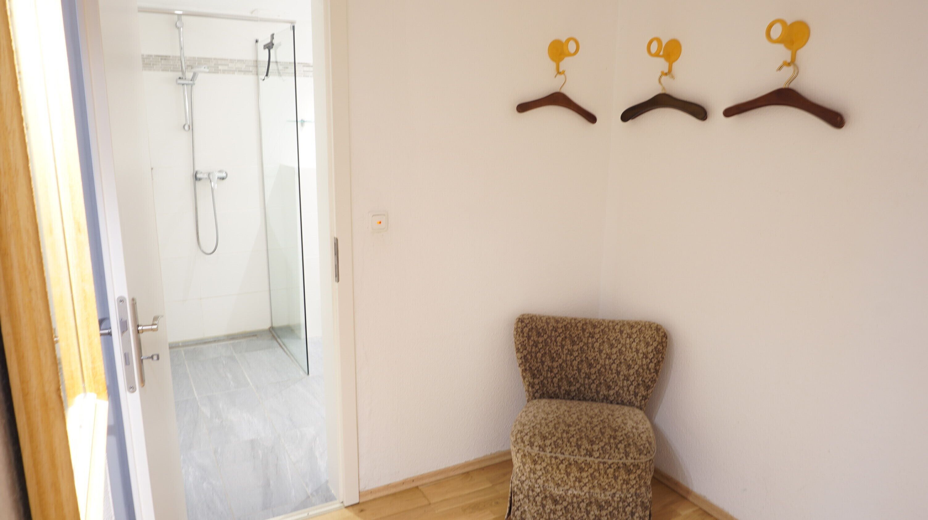 A room with a shower, a chair, and three hangers on the wall.