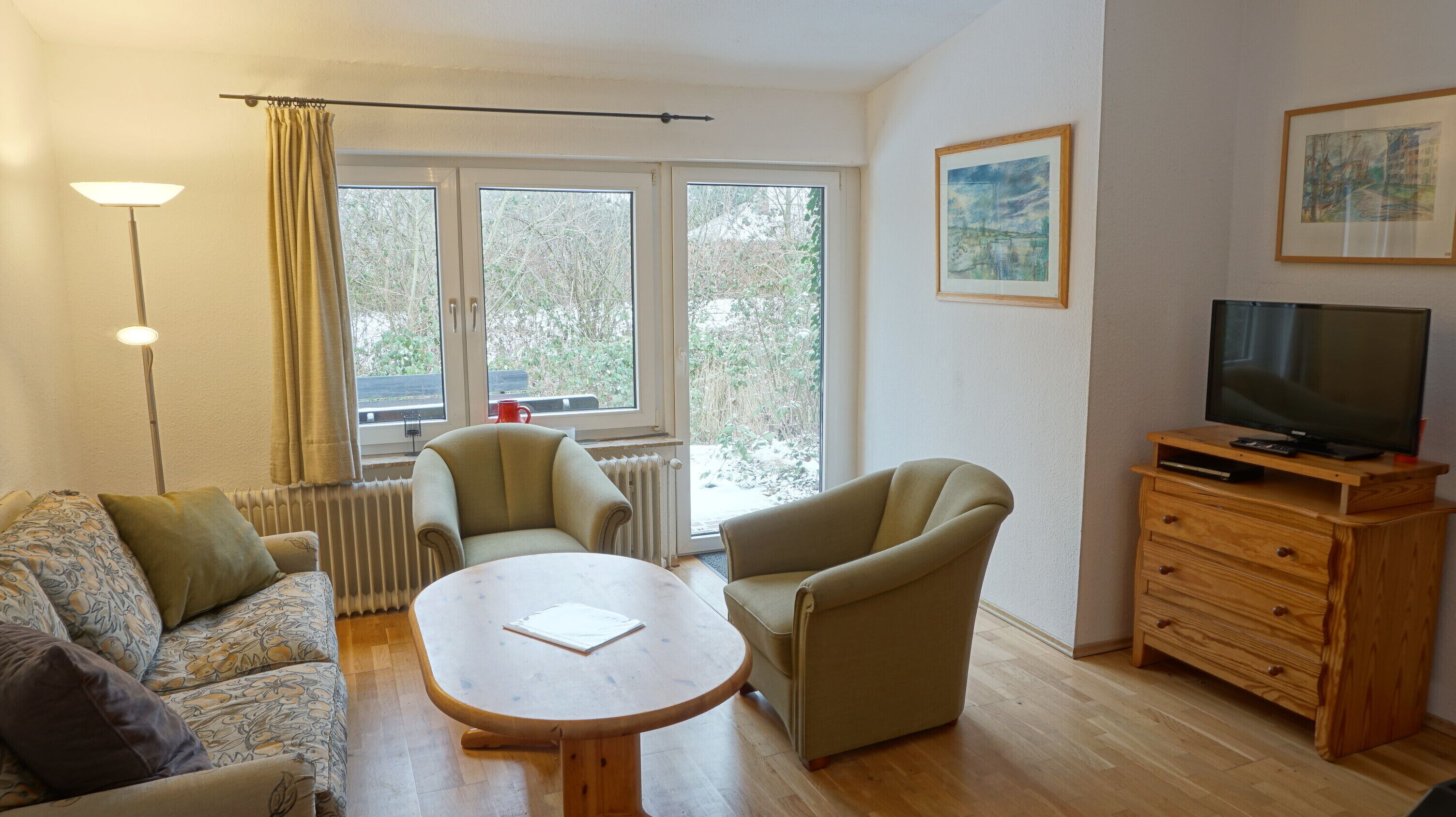 A cozy living room with a sofa, two armchairs, a wooden coffee table, a TV on a wooden cabinet, and a window with a view of trees.