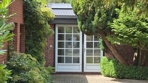 Un edificio de ladrillo con una puerta y ventanas blancas, rodeado de abundante vegetación y árboles.