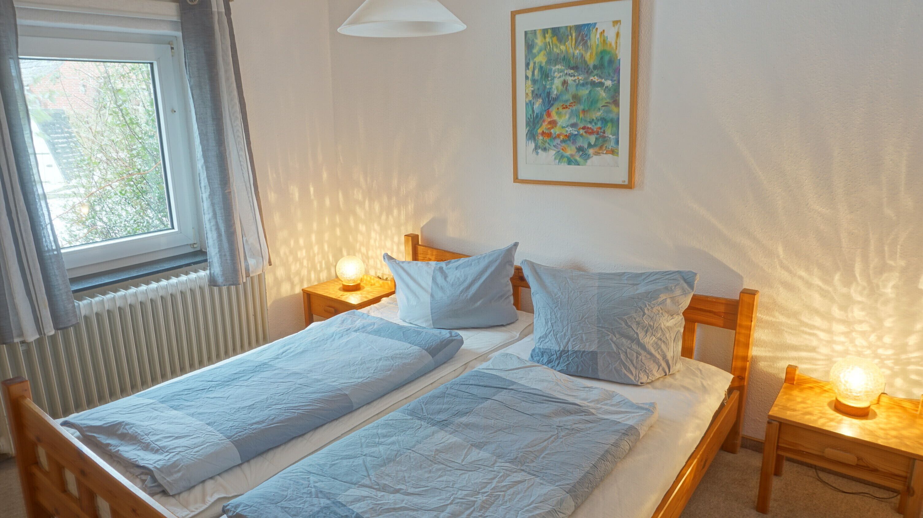 Ein Doppelbett mit zwei Kissen, ein Nachttisch mit Lampe, ein Fenster mit Vorhängen und ein Bild an der Wand.