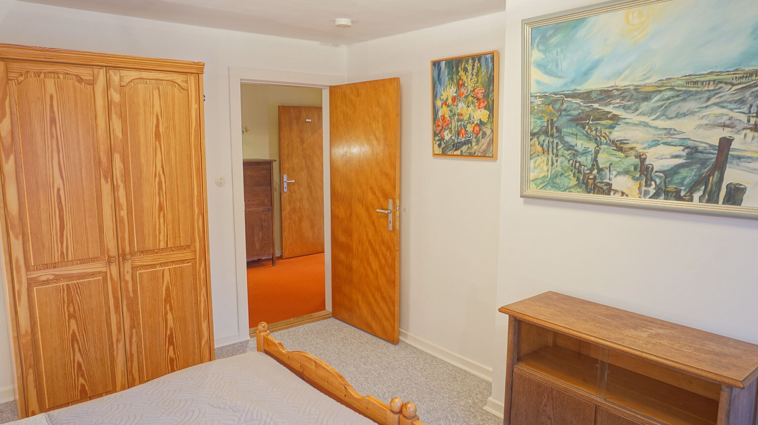 A bedroom with a wooden wardrobe, a bed, a wooden nightstand, and a painting on the wall.