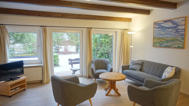 A living room with a sofa, two armchairs, a coffee table, a TV on a stand, and large windows with curtains.