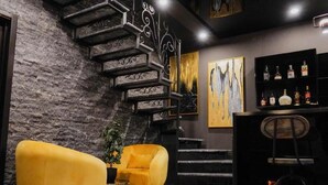 A modern interior with a stone wall, a staircase, two yellow armchairs, a small table with a plant, and a bar area with bottles.