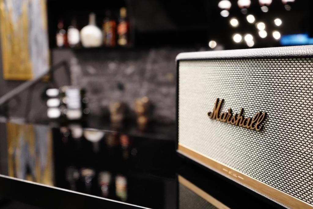 A close-up of a Marshall amplifier with a mesh grille, set against a dark background with blurred bottles and a reflective surface.