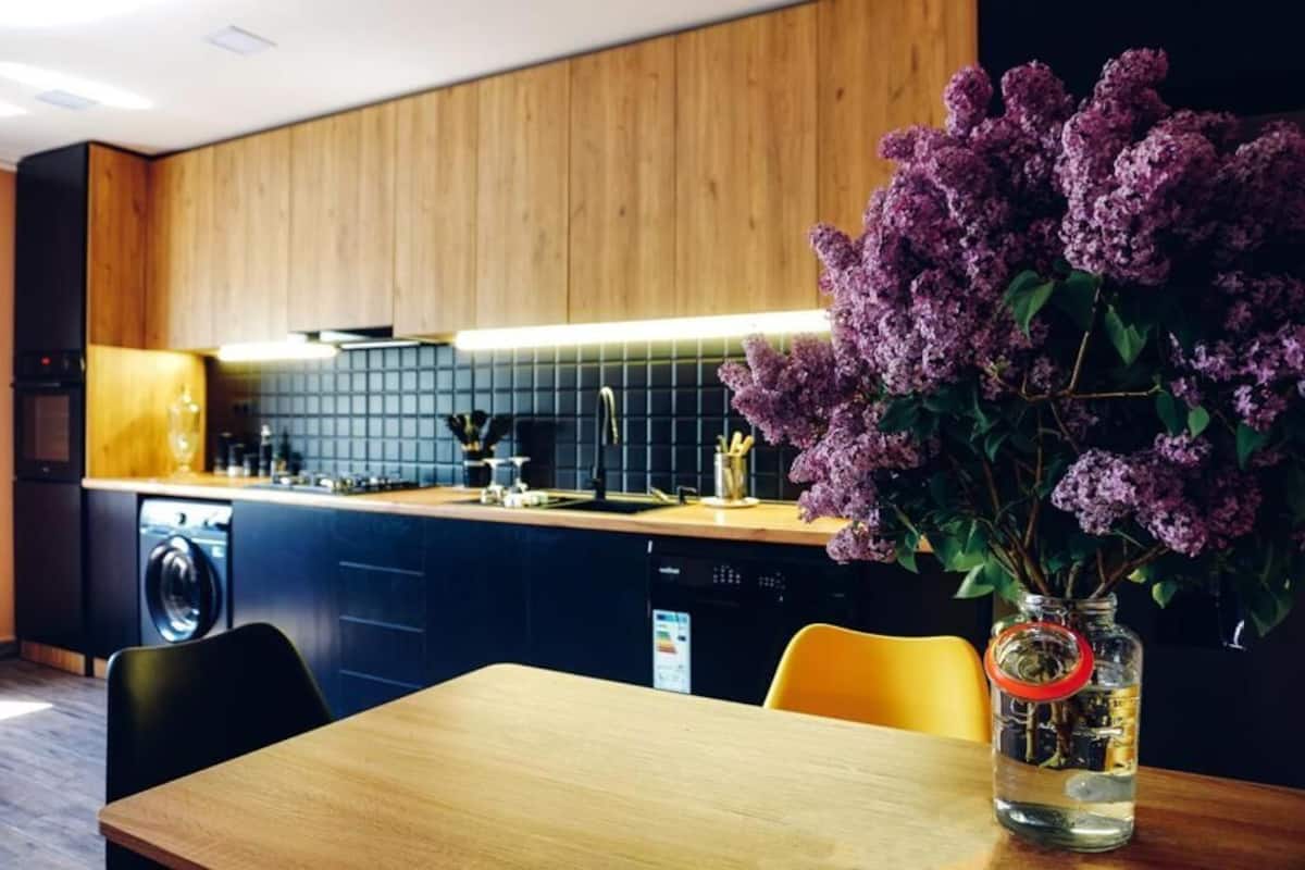 A modern kitchen with wooden cabinets, a black tiled backsplash, and a vase of purple flowers on the counter.