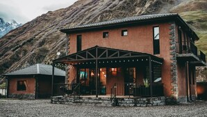 A two-story mountain lodge with a stone facade, a covered porch, and a view of a mountain range.