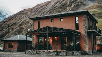 A two-story mountain lodge with a stone facade, a covered porch, and a view of a mountain range.