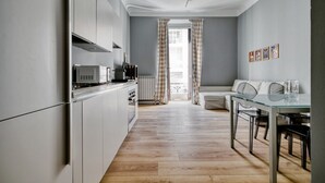 Une cuisine moderne avec des meubles blancs, un coin repas doté d’une table en verre et une vue par une fenêtre ornée de rideaux à carreaux.