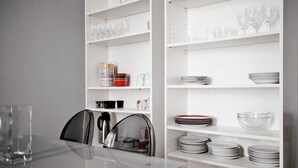 Un coin repas avec une table en verre, des chaises et des étagères blanches où sont disposées des assiettes, des bols et des verres.