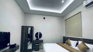 Una habitación de hotel moderna con una televisión de pantalla plana, un escritorio con espejo, una cama con ropa de cama blanca y marrón, y una lámpara de techo.