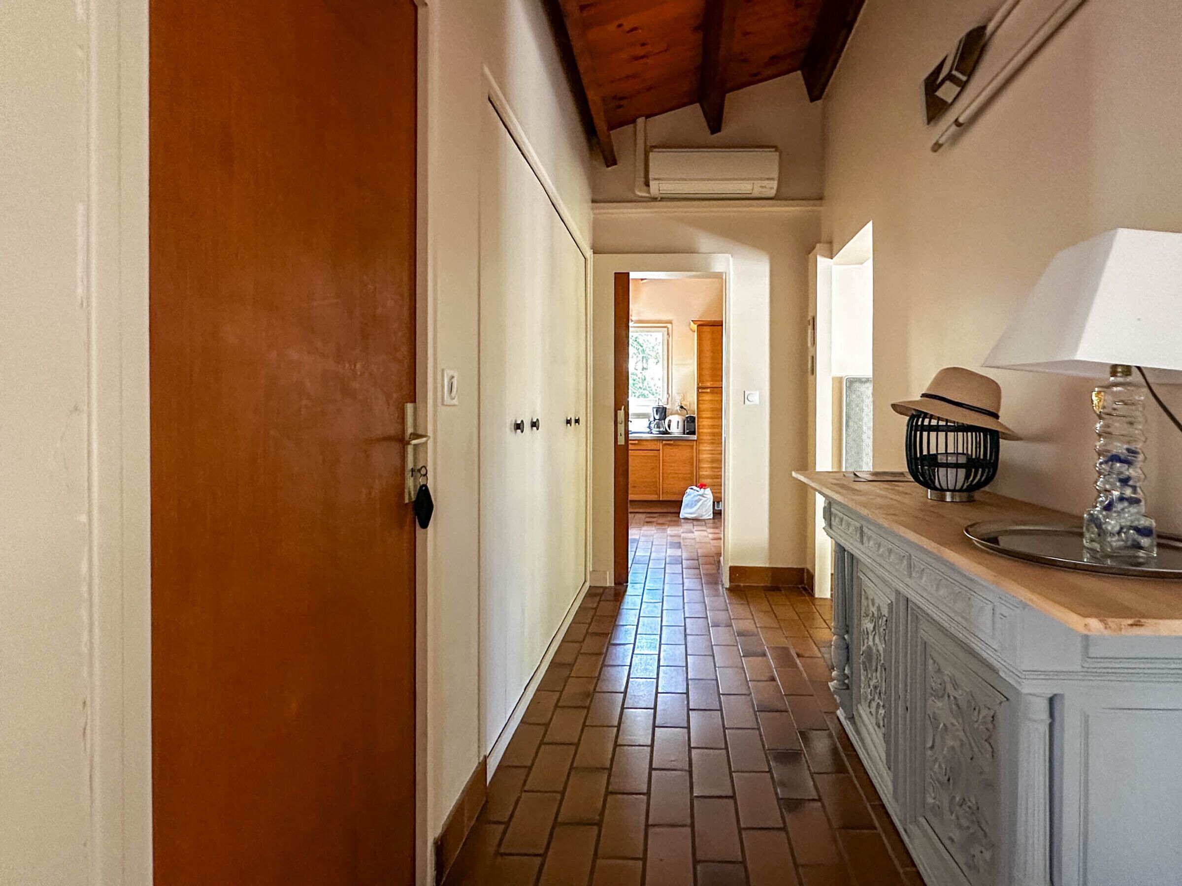 Un pasillo con una puerta de madera, pared blanca, suelo de baldosas, una cocina al fondo y un mueble con una lámpara y objetos decorativos.