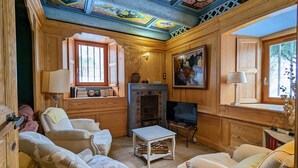 Ein gemütlicher Raum mit Holzwänden, einem Kamin und einer Decke mit farbenfrohen Bildern. Es gibt zwei Sofas und einen kleinen Tisch.