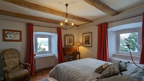 Ein Schlafzimmer mit Bett, Stuhl, Schminktisch, einem Fenster mit Vorhängen, einer Chandelierleuchtre und einem Teppich.