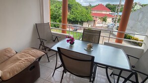 Een balkon met een tafel en stoelen, een bank, en uitzicht op een gebouw met rode daken en begroening.