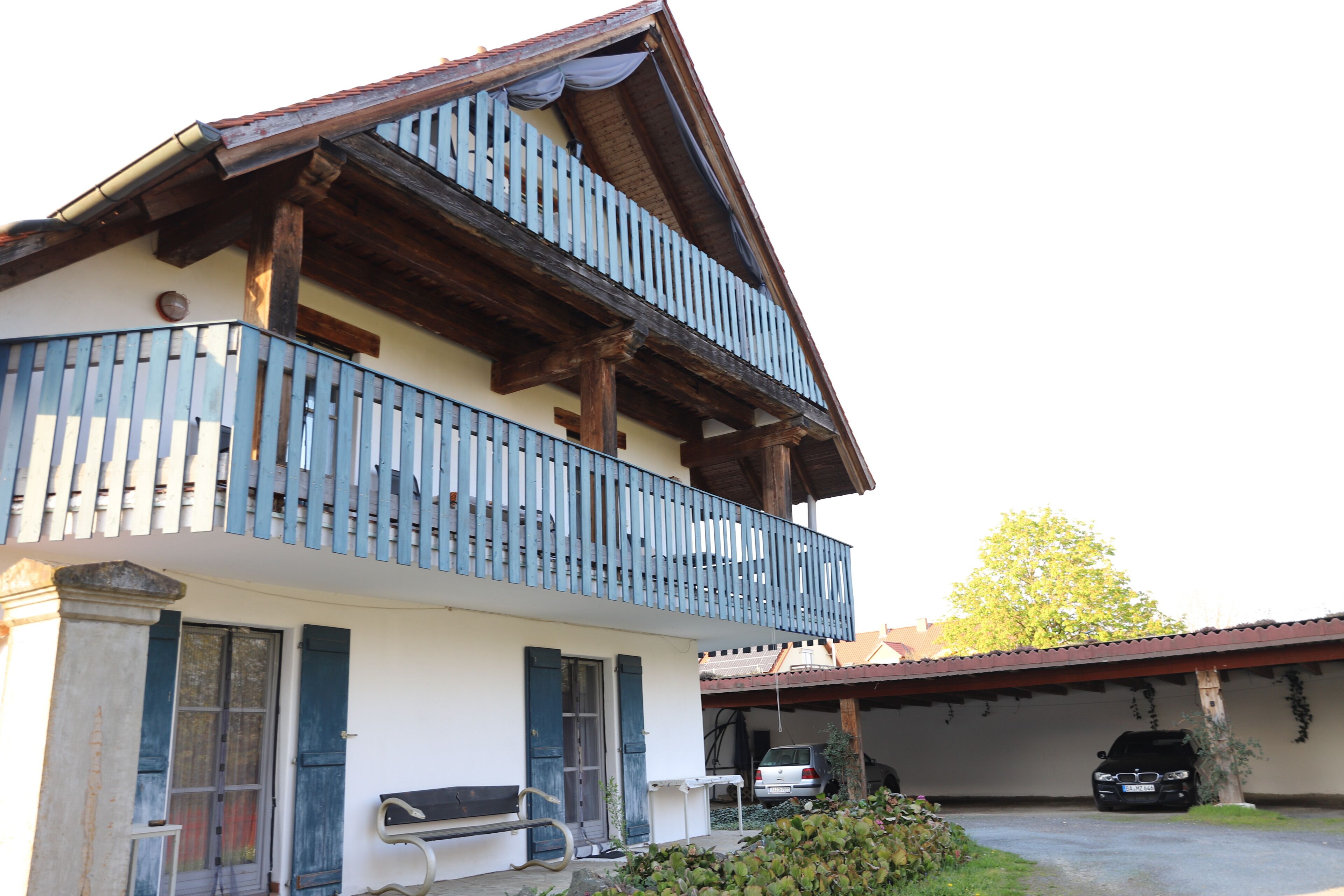 Een gebouw met twee verdiepingen, een balkon, een auto op een overdekte plek en een bank buiten.