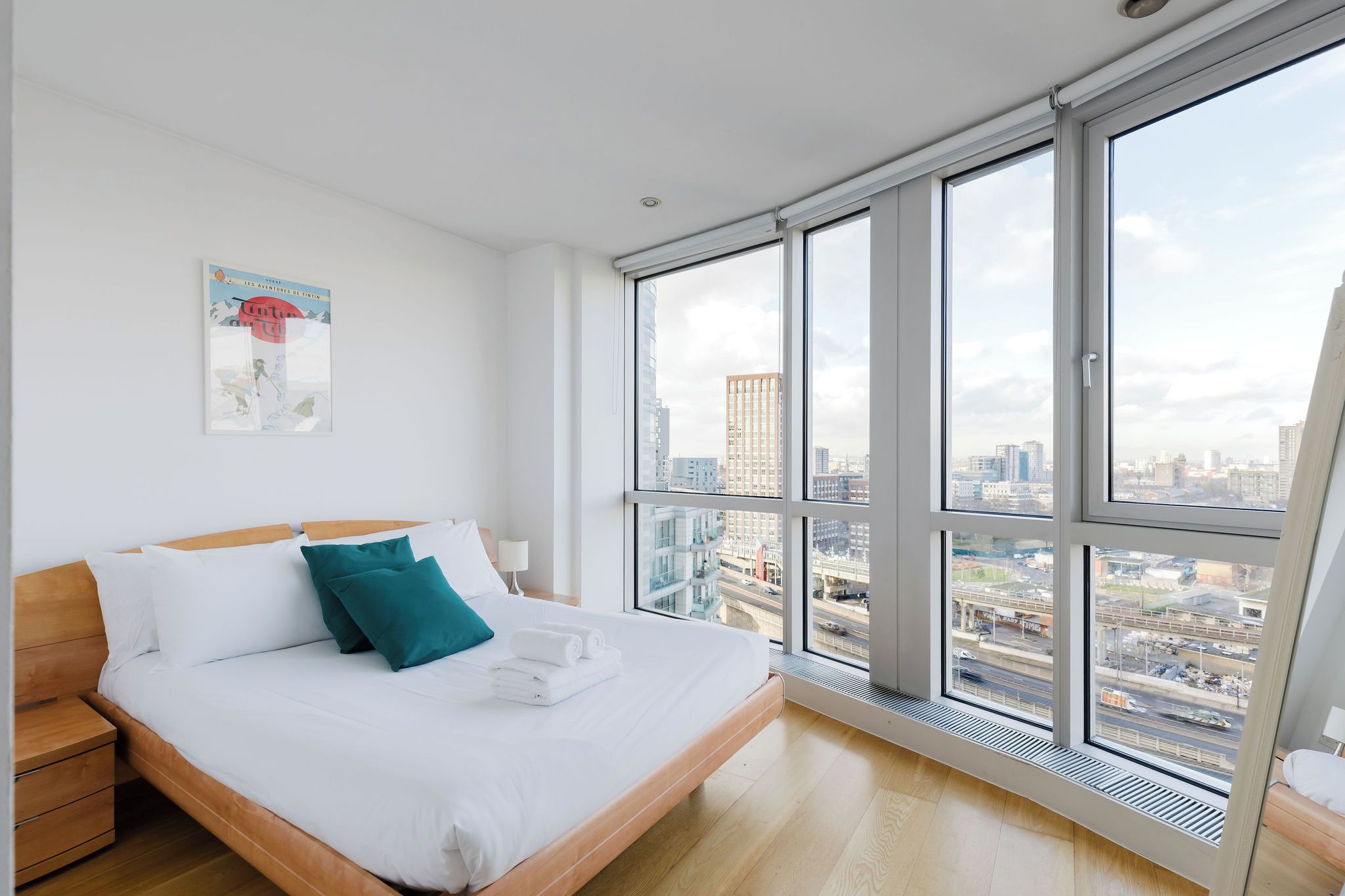 A modern hotel room with a large bed, a nightstand, a window with a city view, and a poster on the wall.