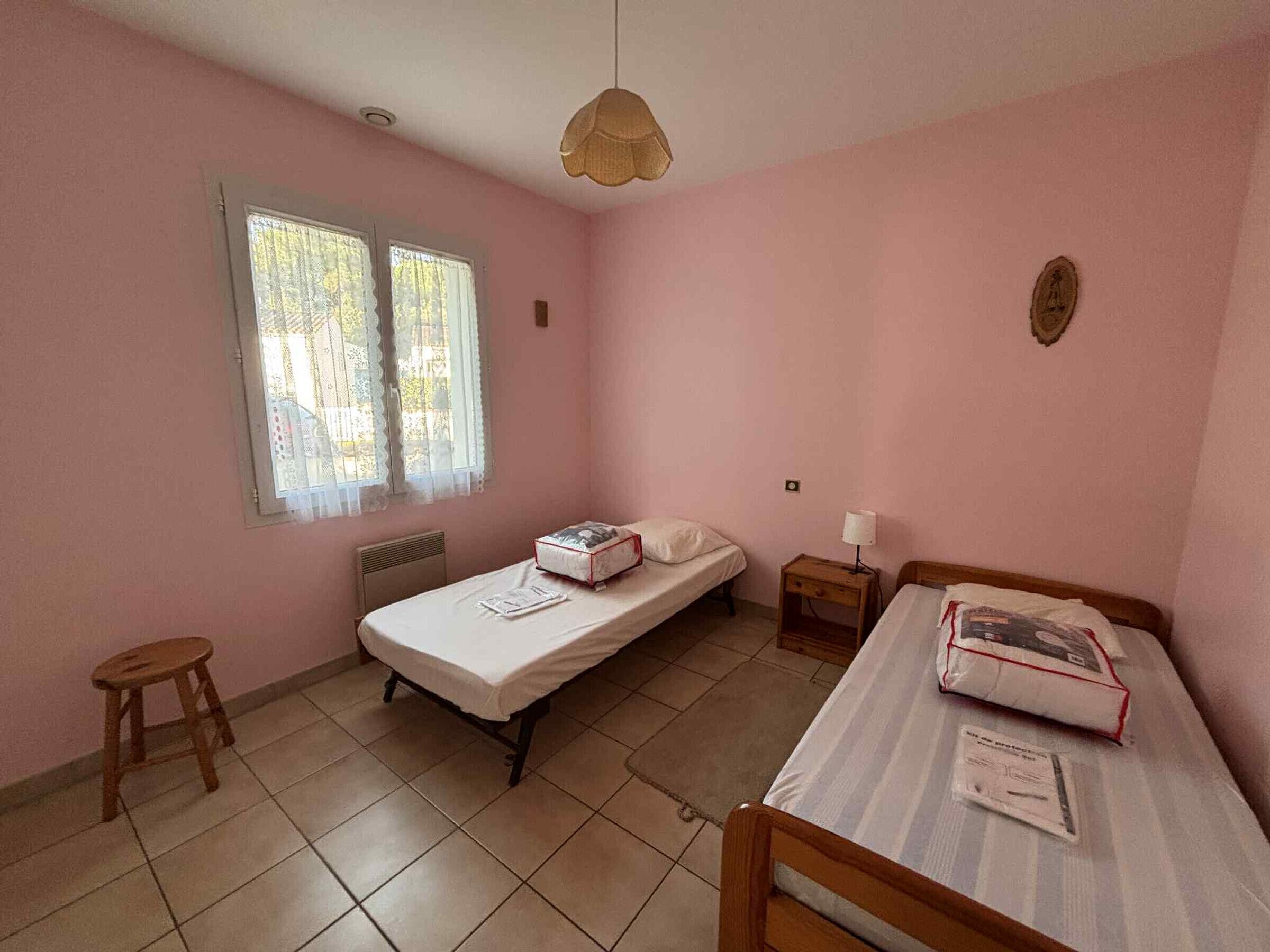Una piccola camera da letto con pareti rosa, due letti singoli, uno sgabello e una finestra con vista sugli alberi.