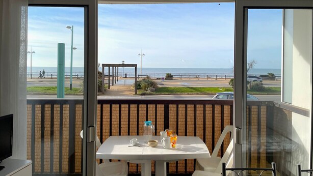 Varanda com mesa posta para dois, com vista para uma praia, uma passarela para pedestres e carros estacionados.