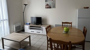 A compact living space with a dining table, chairs, a television on a cabinet, a refrigerator, and a window with curtains. - Central Apartment for 4 - Parking Included (Royan)