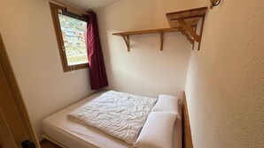 Una camera piccola con un letto singolo, una testiera in legno, una finestra con tende e uno scaffale in legno sopra il letto.