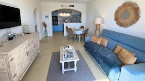 Un soggiorno con divano, tavolino e televisore. Sullo sfondo, una cucina con mobili blu e un'area pranzo.