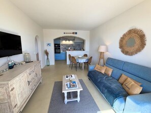 A living room with a sofa, coffee table, and TV. There is a kitchen in the background with blue cabinets and a dining area. - 2-Bedroom Apartment with Air Conditioning and WIFI (Port Grimaud)
