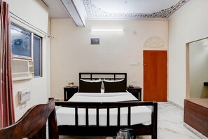 Une chambre à coucher comprenant un lit, une tête de lit en bois, une table de chevet, une commode, une fenêtre avec des rideaux, un climatiseur fixé au mur et une porte ornée d’un motif décoratif.